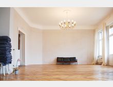 Minimalistisch eingerichteter heller Raum mit Parkettboden, einer einfachen Sitzbank, einer hängenden goldfarbenen Kronleuchterlampe und großen Fenstern mit weißen Vorhängen. Links im Raum sind gestapelte Decken und eine Topfpflanze sichtbar.