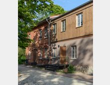 Backstein-Gebäude mit einer holzverkleideten Erweiterung und großen Fenstern, umgeben von Bäumen. Vor dem Gebäude befinden sich Metalltreppen mit Geländern sowie ein gepflasterter Weg.