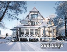 Fachwerkhaus im Winter mit verschneiter Umgebung, das auf Stelzen am Ufer steht. Im Vordergrund sind schneebedeckte Wege und Bäume, während das Gebäude mit großen Fenstern und einem roten Ziegeldach gestaltet ist.