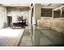 Industrieinspirierter Raum mit freigelegten Backsteinwänden, einem historischen schwarzen Auto, Holzboden und einer Fahrradecke. An der Wand hängen gerahmte Bilder, während große Fenster und hölzerne Elemente die Einrichtung ergänzen.