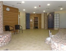 Wellnessbereich mit gefliesten Wänden und bräunlichem Holzpaneel an den Seiten. Zu sehen sind runde Sitzgelegenheiten mit Mosaikfliesen, zwei Stühle aus dunklem Geflecht sowie eine Sauna mit Glastür im Hintergrund. Deckenstrahler und beige Fliesenboden prägen den Raum.