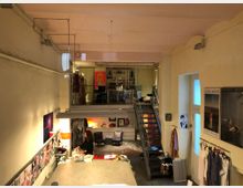 Ein heller, zweistöckiger Arbeitsraum mit hohen Decken. Im Vordergrund ein großer Arbeitstisch mit Nähmaterialien, hinten eine Metalltreppe zu einer Galerieebene mit Regalen und Kleiderständern. Wände mit Postern und Bildern dekoriert, im Hintergrund eine Sitzgruppe und eine Figurine mit Kleidern.