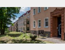 Mehrfamilienhaus in einer Wohnsiedlung mit hellen Fassaden, roten Ziegeldetails und mehreren Balkons; im Vordergrund eine kleine Grünfläche mit Bäumen und niedrigen Pflanzen.