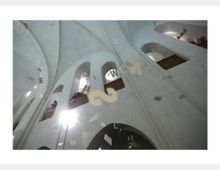 Innenaufnahme einer hellen Kirche mit hohen, gewölbten Decken und schmalen Rundbogenfenstern. Personen stehen auf einer oberen Galerie, während Buchstaben oder Symbole im Raum zu schweben scheinen.