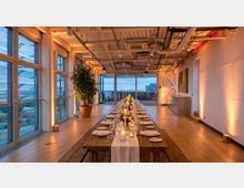 Modern loft-style event space with floor-to-ceiling windows overlooking a city, wood floors, and an exposed industrial ceiling with ducts and lighting. A long banquet table is set with plates, glassware, candles, and floral arrangements, with a large potted plant and wall art along the side.