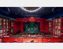 Innenraum des Wintergarten-Varieté-Theaters mit rotem Bühnenvorhang, Bühne mit Musikinstrumenten und einer geschwungenen Metallkulisse. Der Raum ist dekoriert mit beleuchteten Postern an den Wänden und einer dunkelblauen Decke mit Sternenmotiven.