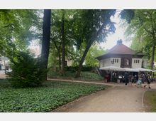 Ein kleiner Park mit dichtem Baumbestand und bodenbedeckendem Efeu. Im Hintergrund ein kleines Gebäude mit Fachwerkdetails und einem roten Ziegeldach, vor dem mehrere weiße Sonnenschirme und Menschen zu sehen sind.
