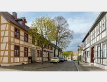 Straße mit Kopfsteinpflaster in einem historischen Viertel, gesäumt von Fachwerkhäusern in hellen Farben mit rotbraunen und grauen Akzenten. Im Vordergrund stehen Bäume mit frischem Frühlingslaub und eine Reihe geparkter Autos.