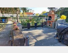 Gepflasterte Garten- und Terrassenfläche mit Pflanzkübeln und Blumenbeet entlang eines Gehwegs, eingefasst von niedrigen Steinmauern und einem Metallzaun. Rechts steht ein gemauerter Grillkamin neben abgedeckten Gartenmöbeln; im Hintergrund sind Wohnhäuser und Bäume zu sehen.