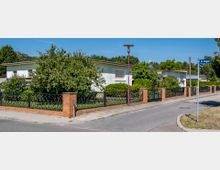 Straßenszene in einem Wohngebiet mit einstöckigen weißen Häusern, Vorgärten und Zäunen aus schwarzem Metall mit gemauerten Pfosten. Im Vordergrund ein Gehweg, eine Straßenlaterne und ein Straßenschild mit der Aufschrift „Im Bogen“.