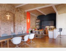 Offener Wohn- und Essbereich in einem Loft mit Backsteinwänden, einem Gewölbedeckenstil und Holzboden. Der Raum umfasst einen Essbereich mit modernem Tisch und weißen Stühlen, eine Küche mit schwarzen Einbauschränken, Edelstahlgeräten und einem freistehenden Küchenblock, sowie Akzente in leuchtendem Orange.