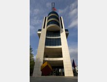 Das Bild zeigt das Willy-Brandt-Haus, ein modernes Gebäude mit heller Fassade, symmetrischen Strukturen und einer auffälligen runden, verglasten Mittelpartie. Vor dem Eingang steht ein schräg platzierter, rot-gelber Würfel, und links daneben wehen Fahnen.