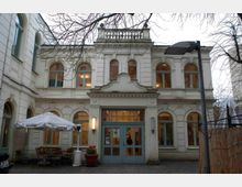 Historische Gebäudeansicht mit heller Putzfassade und bogenförmigen Fenstern, zentral ein zweiflügeliger Eingang mit blauen Türen. Im Vordergrund ein kleiner Außenbereich mit Tischen, Stühlen und einem Wetterschirm.