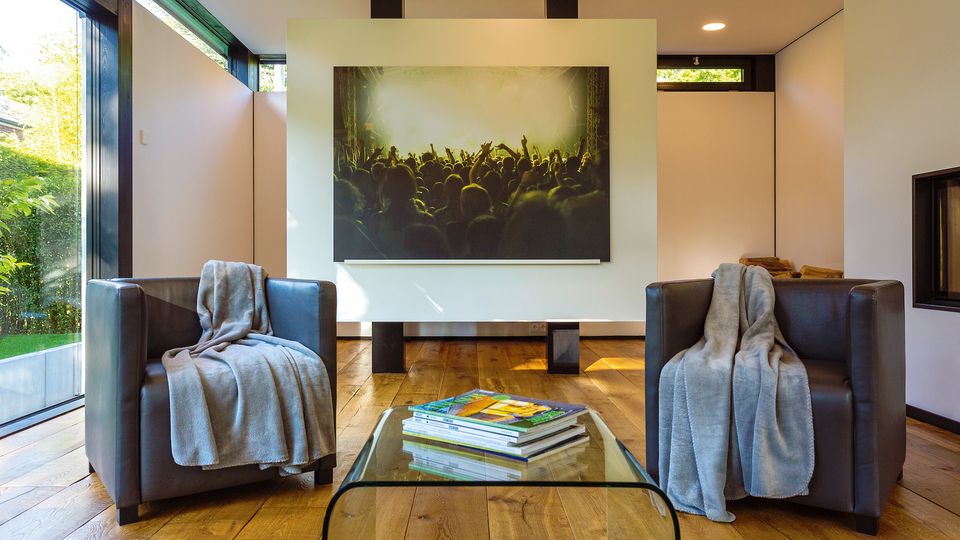 Moderner Wohnraum mit Holzfußboden, zwei schwarzen Ledersesseln und einem niedrigen Glastisch mit Zeitschriften. Große bodentiefe Fenster und eine zentrale Wand mit großformatigem Bild prägen den Raum, daneben ist ein eingebauter Kamin zu sehen.