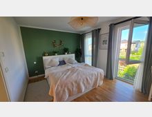 Modernes Schlafzimmer mit Doppelbett vor einer dunkelgrünen Akzentwand, hellen Betttextilien und Holzfußboden. Rechts sorgen große bodentiefe Fenster mit grauen Vorhängen und geöffnetem Flügel für viel Tageslicht und Blick ins Wohngebiet mit Garten.