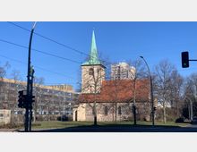 Historische Backsteinkirche mit rotem Satteldach und einem Turm mit grünem Spitzhelm, umgeben von kahlen Bäumen, im Hintergrund moderne Wohn- und Bürogebäude. Im Vordergrund eine Straßenkreuzung mit Ampeln und Straßenlaternen.