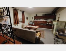 Wohnzimmer im Retro-Stil mit Möbeln und Dekoration aus den 1970er Jahren. Zu sehen sind eine hellgraue Couch, ein Couchtisch, ein dunkler Holzschrank mit Glasvitrine, ein alter Röhrenfernseher und ein grüner Kachelofen; im Vordergrund ein Kinderwagen, an der Wand braune Vorhänge.