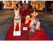 Ausstellung mit antiken Kinderfahrzeugen, darunter ein bemaltes Holzpferd, ein Holzhund und ein Kinderroller im Retro-Stil, auf einem roten Podest präsentiert. Im Hintergrund sind Informationstafeln mit Texten und Bildern sowie eine abgesperrte Ausstellungsfläche sichtbar.