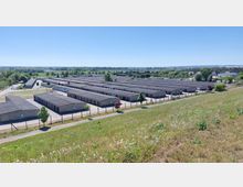 Weitläufige Anlage mit mehreren rechteckigen, flachen Lagerhallen aus Beton, umgeben von einem Zaun und einzelnen Bäumen. Im Hintergrund sind grüne Felder, Bäume und kleinere Gebäude einer Ortschaft erkennbar.