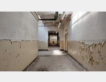 Ein verlassener Gang in einem heruntergekommenen Gebäude mit abblätternder Farbe an den Wänden, freiliegenden Rohren an der Decke und Schutt auf dem Boden. Auf der rechten Seite ist eine Tür mit der Aufschrift „Fremd“.