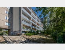 Mehrstöckiges Wohngebäude in Betonbauweise mit Balkonen, die mit Blumen und Pflanzen dekoriert sind. Im Vordergrund eine Treppe, die zu einem leicht bewachsenen Hang mit Bäumen führt, umgeben von städtischem Grün.