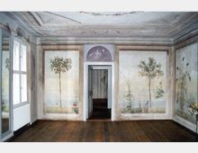 Historischer Innenraum mit bemalten Wanddekorationen, die Bäume und Pflanzen in einer natürlichen Landschaft darstellen. Der Raum hat Holzböden, eine bemalte Decke mit Ornamenten und eine Tür, die zu einem benachbarten dekorierten Raum führt.
