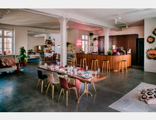 Großzügiger offener Wohnraum mit modernem Design, bestehend aus einem Essbereich mit gedecktem Holztisch, einer stilvollen Küche mit Holztheke und Barhockern sowie einer gemütlichen Sitzecke mit Sofas. Die Einrichtung umfasst Zimmerpflanzen, dekorative Regale, bunte Akzente und helle Fenster, die den Raum mit natürlichem Licht durchfluten.