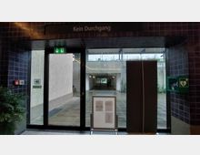 Ein verglaster Eingang mit der Aufschrift "Kein Durchgang", dahinter ein überdachter Durchgang mit Sicht in die Ferne. Links befindet sich ein Feueralarm und rechts ein Defibrillator-Kasten an gekachelten Wänden.