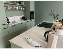 Moderne Küche mit grünen Schränken, einer weißen Arbeitsplatte und einem zentralen Küchenblock mit integriertem Herd. Regale mit Vorratsgläsern, ein Waschbecken mit Wasserhahn und diverse Küchengeräte sind sichtbar, zusammen mit einer Vase mit Blumen als Dekoration.