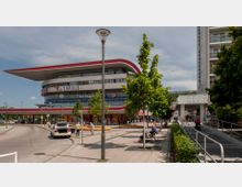 Modernes Einkaufszentrum mit geschwungener, futuristischer Dachkonstruktion und großen Glasfassaden. Im Vordergrund Sitzbänke, Gehweg, einige Menschen sowie kleinere Geschäfte, darunter ein Piercing- und Tattoo-Studio; im Hintergrund Parkplätze und ein Wohngebäude.