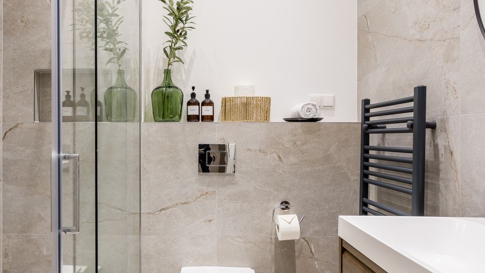 Modernes Badezimmer mit begehbarer Glasdusche links, wandhängendem WC in der Mitte und Waschtisch mit Holzfront rechts. Helle Steinfliesen, ein schwarzer Handtuchheizkörper und eine Ablage mit grünen Glasvasen und Pflegeflaschen prägen den Raum.