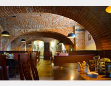 Gemütliches Restaurant mit Backsteinwänden und Gewölbedecke. Holztische und -stühle sind in Reihen angeordnet, mit modernen Hängelampen, die den Raum beleuchten; im Hintergrund sind Edelstahl-Brauanlagen sichtbar.