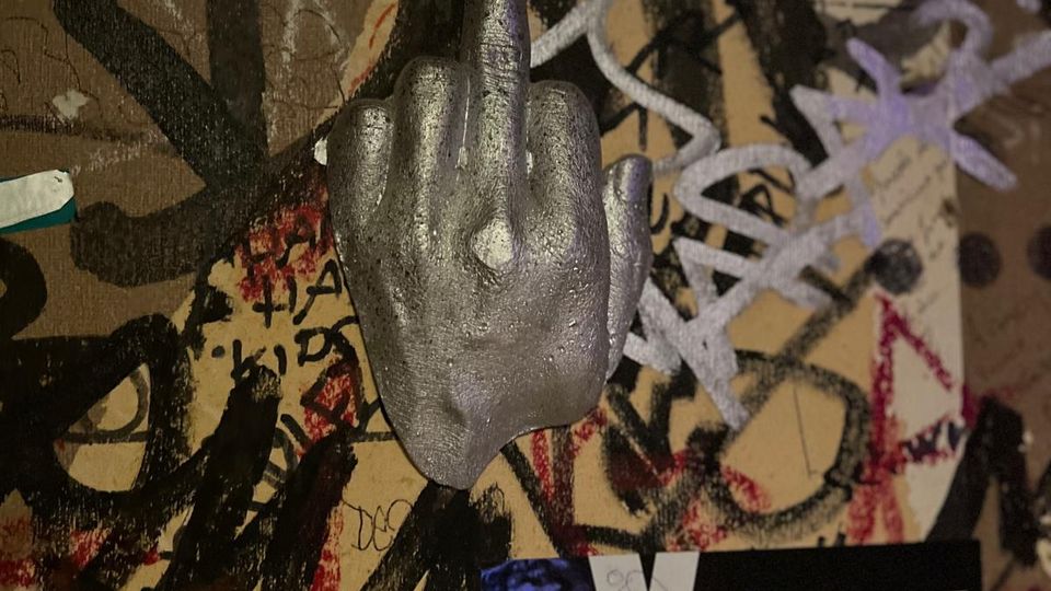 Nahaufnahme einer mit Graffiti, Kritzeleien und Aufklebern bedeckten Wand, in deren Mitte eine silberne Hand-Skulptur montiert ist. Unten ist ein rechteckiger Sticker mit der Aufschrift „MC SÄLZBROT“ zu sehen.