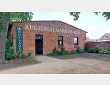 Backsteingebäude mit der Beschriftung „REGIONALMARKTHALLE“ auf der Frontseite, darunter ein Eingang und zwei Fenster mit Efeu bewachsen; ein grünes Banner links trägt die Aufschrift „Hofladen“. Im Vordergrund ein unbefestigter Weg, umgeben von Pflanzen und Bäumen.