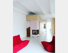 Modernes Wohnzimmer mit weißem Boden und Wänden, einer rosa Kaminverkleidung mit Holzelementen, einem Stapel Brennholz daneben und zwei roten Lounge-Sesseln im Vordergrund. Im Hintergrund befindet sich eine offene Küche mit hellen Holzmöbeln.