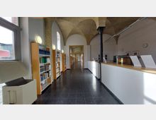 Ein heller Flur in einer Bibliothek mit Regalen voller Bücher auf der linken Seite und einer Empfangstheke auf der rechten Seite. Die Architektur zeigt gewölbte Decken aus Ziegelstein und schwarze Metallstützen; am Ende des Flurs befinden sich Türen.