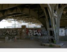 Unterseite einer Stahlbrücke mit massiven, genieteten Trägern, die von gemauerten Pfeilern gestützt wird. Die Wände sind mit zahlreichen Graffiti bedeckt, und der Boden darunter ist unbefestigt mit vereinzeltem Schutt.