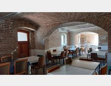 Raum mit Gewölbedecke aus unverputztem Ziegelstein, mehreren rechteckigen Tischen mit Kunststoffdecken und verschiedenen Stühlen. Durch Fenster fällt Tageslicht herein, und an den Seiten sind hölzerne Türen und weiße Wände im unteren Bereich sichtbar.