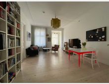 Langer Wohnraum mit hellem Holzboden, links ein Bücherregal und ein Sofa, rechts ein Esstisch mit weißen Stühlen und rotem Gestell. Im Hintergrund stehen ein Fernseher mit Sideboard, ein Sessel und ein Fenster mit weißen Vorhängen, das viel Tageslicht hereinlässt.