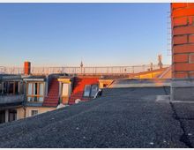 Blick über die Dächer einer Stadt mit roten Ziegeldächern und großen Fenstern, im Hintergrund ist der Berliner Fernsehturm vor einem klaren Himmel bei Sonnenuntergang zu sehen. Im Vordergrund ist ein flaches Dach mit einer Backsteinwand rechts im Bild.