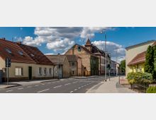 Straßenszene in einer Kleinstadt mit mehreren Gebäuden im typischen Stil, darunter Wohnhäuser mit Ziegeldächern und ein historisches Gebäude mit dekorativem Giebel. Die Straße hat Fußwege, Straßenlaternen und eine Ampel, die auf Grün steht, bei klarem Himmel mit wenigen Wolken.