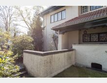 Außenansicht eines Wohnhauses mit hell verputzten Wänden, rotem Ziegeldach und dekorativen Fenstergittern. Im Vordergrund befindet sich eine niedrige Mauer mit Treppe, umgeben von Gartenbepflanzung mit Büschen und Bäumen.