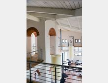 Heller Innenraum eines Gebäudes mit sichtbaren weißen Holzbalken an der Decke, Ziegelbögen in den Wänden und offenen Galerien aus Metall und Holz. Der Raum ist modern gestaltet mit mehreren Tischen und Stühlen im Erdgeschoss und großen, zylindrischen Hängelampen.