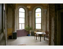 Innenraum eines alten, verfallenen Gebäudes mit hohen, bogenförmigen Fenstern mit buntem Glas und dekorativen Säulen. Der Raum enthält einfache Möbel, darunter einen Tisch, Stühle und einen kleinen Schrank sowie eine Topfpflanze.