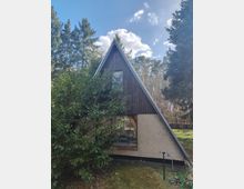 Holzhütte in A‑Rahmen-Architektur mit steilem Satteldach und kleinem Fenster im Giebel, teilweise von einem großen Strauch verdeckt. Das Gebäude steht auf einer Waldlichtung, umgeben von hohen Nadelbäumen, mit Rasenfläche und Gartenschlauch im Vordergrund.