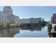 Historisches Gebäude mit Kuppel neben einem Kanal, über den eine steinerne Brücke führt. Im Hintergrund sind moderne und klassische Gebäude sowie ein klarer Himmel sichtbar.