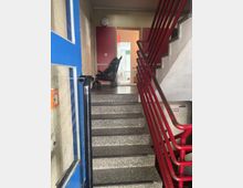 Treppenhaus mit grauen Stufen und einem roten Geländer, das nach oben führt. Im Hintergrund steht ein zusammengeklappter schwarzer Kinderwagen, und durch eine offene Tür ist ein heller Raum mit einem grünen Lampenschirm sichtbar.