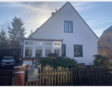 Ein weißes Einfamilienhaus mit Satteldach, einem Wintergarten mit großen Fenstern im Vordergrund und einem hölzernen Zaun, der das Grundstück umgibt. Im Garten befinden sich grüner Strauchbewuchs, und links neben dem Haus steht ein überdachter Carport mit einem geparkten Auto.