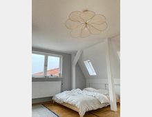 Schlafzimmer in einem Dachgeschoss mit minimalistischem Stil, einem niedrigen Holzbett mit weißer Bettwäsche, großen Fenstern mit Dachblick und einem dekorativen Lampenschirm in Blütenform an der Decke.