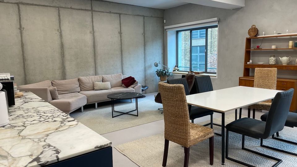 Open-plan modern living and dining area with exposed concrete walls and ceiling lights, featuring a sectional sofa around a round coffee table and a white dining table with mixed chairs on a rug. A large window, marble-look kitchen counter edge, and a wooden shelving unit with decor complete the space.
